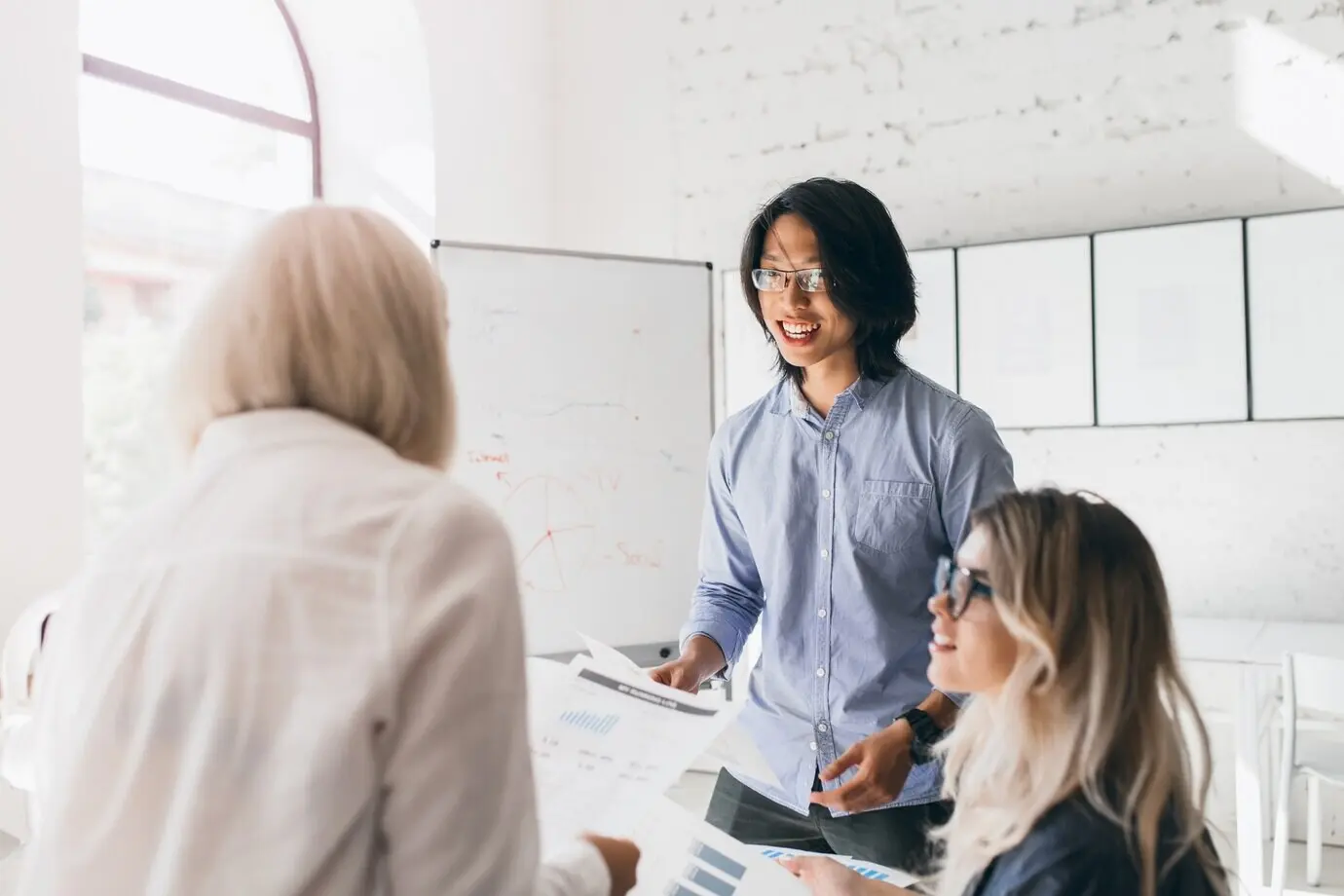 Porträt von hinten einer blonden Frau in einem stylischen Hemd, die Dokumente liest, während ein asiatischer Mann ihr etwas erzählt.
